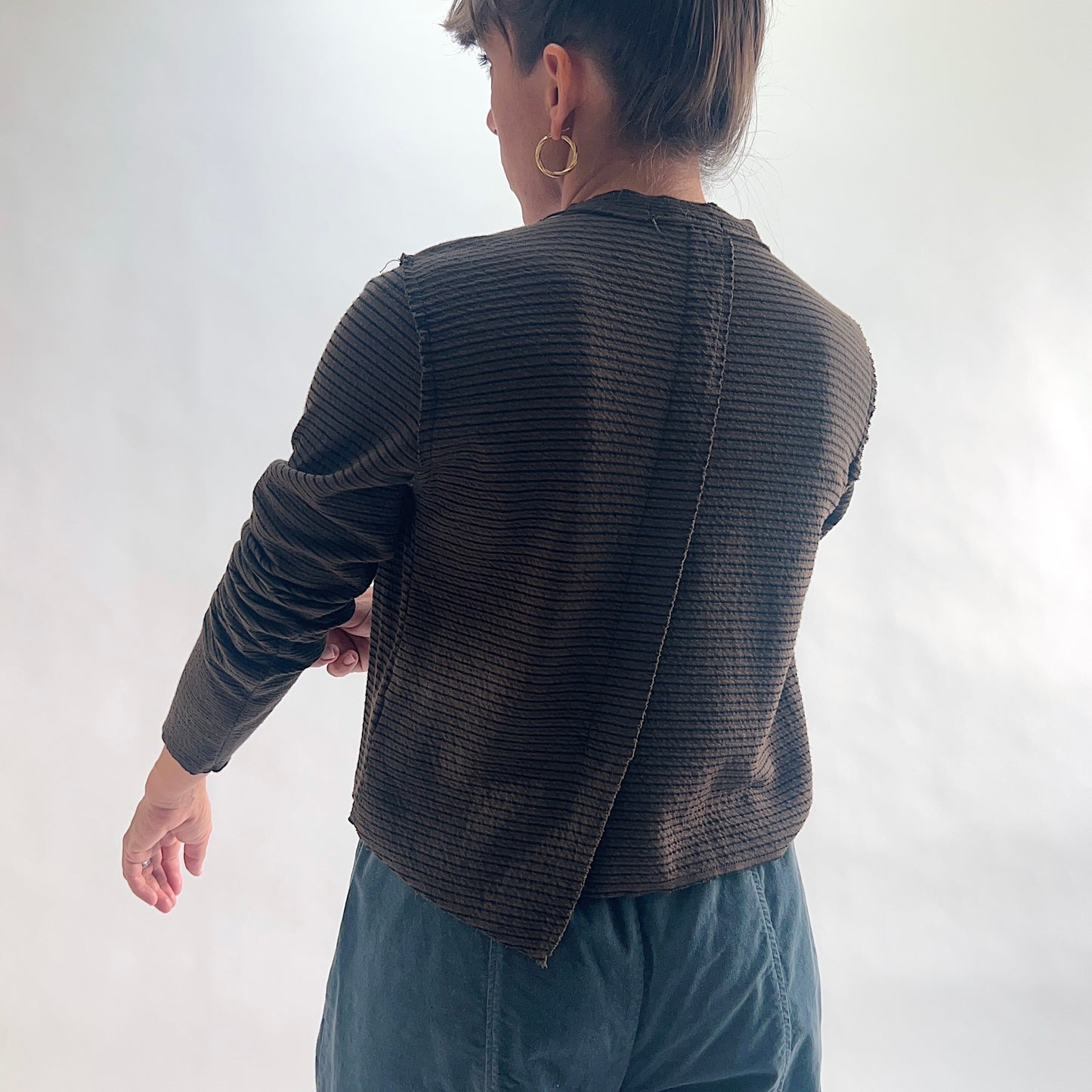 Back view of model wearing a chocolate and black fine tonal stripe cropped cardigan with long sleeves and asymmetrical hem, arm extended, showing the center back seam and crinkle-texture fabric, styled with teal wide-leg pants against a white background.
