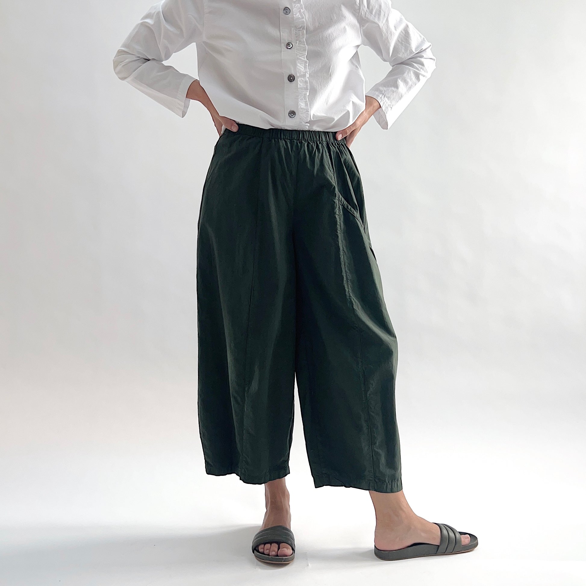 Full front view of model wearing Eleven Stitch konbu one pocket pant with hands on hips, showing elastic waistband, wide curved-seam cropped leg, large curved side pocket, and deep forest green color, styled with white long sleeve button front shirt and dark slide sandals