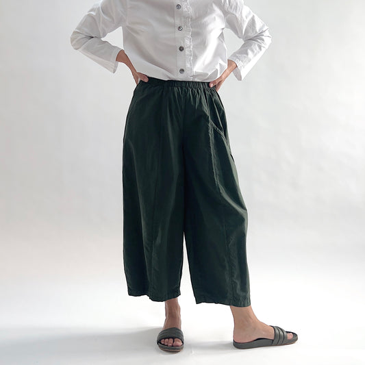 Full front view of model wearing Eleven Stitch konbu one pocket pant with hands on hips, showing elastic waistband, wide curved-seam cropped leg, large curved side pocket, and deep forest green color, styled with white long sleeve button front shirt and dark slide sandals