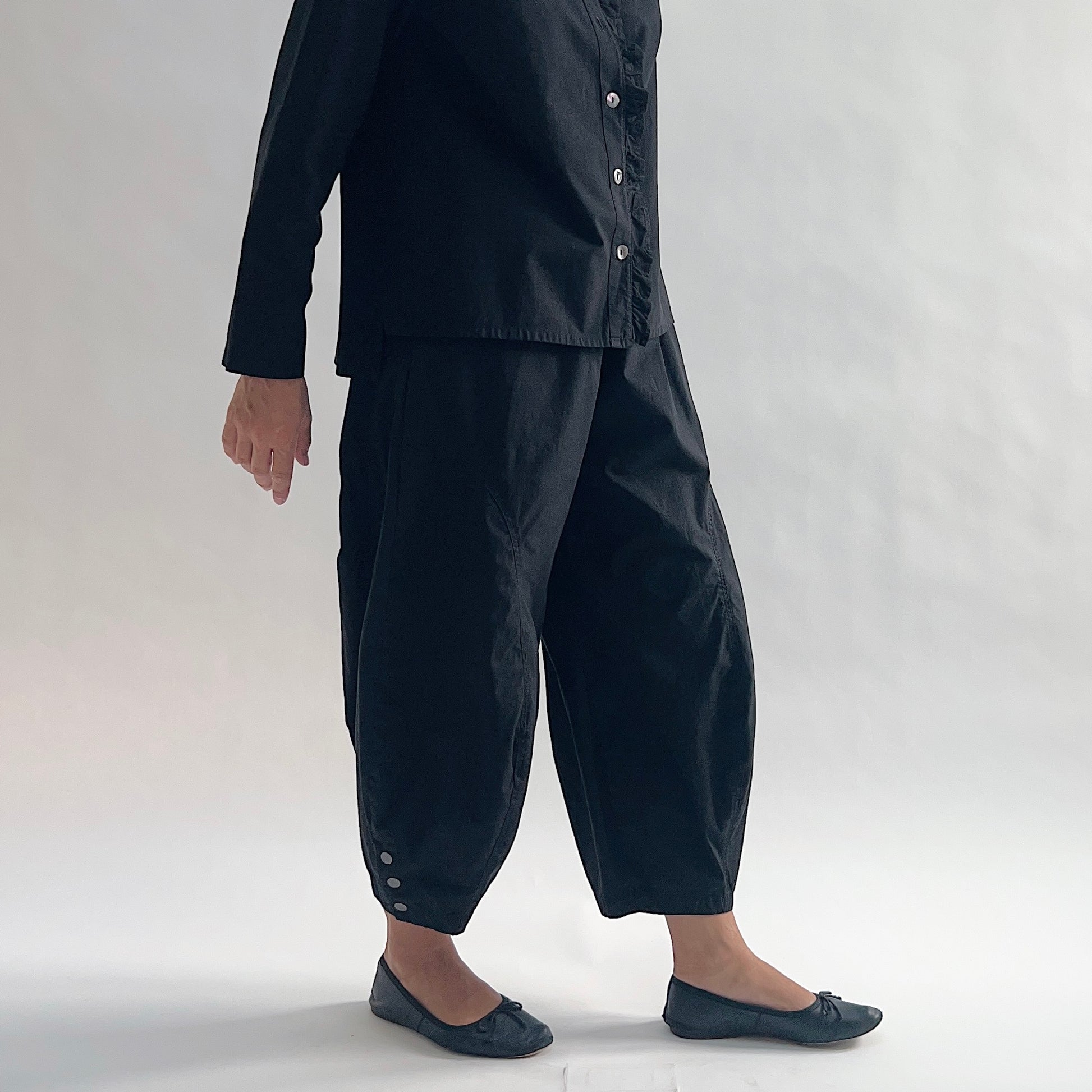 Side view of model with hand on hip wearing Eleven Stitch black snap doreen pant, showing elastic waistband, wide balloon leg tapering to snap hem at ankle, side seam pocket, snap buttons at ankle, and black color, styled with matching black blossom collar shirt and black ballet flats