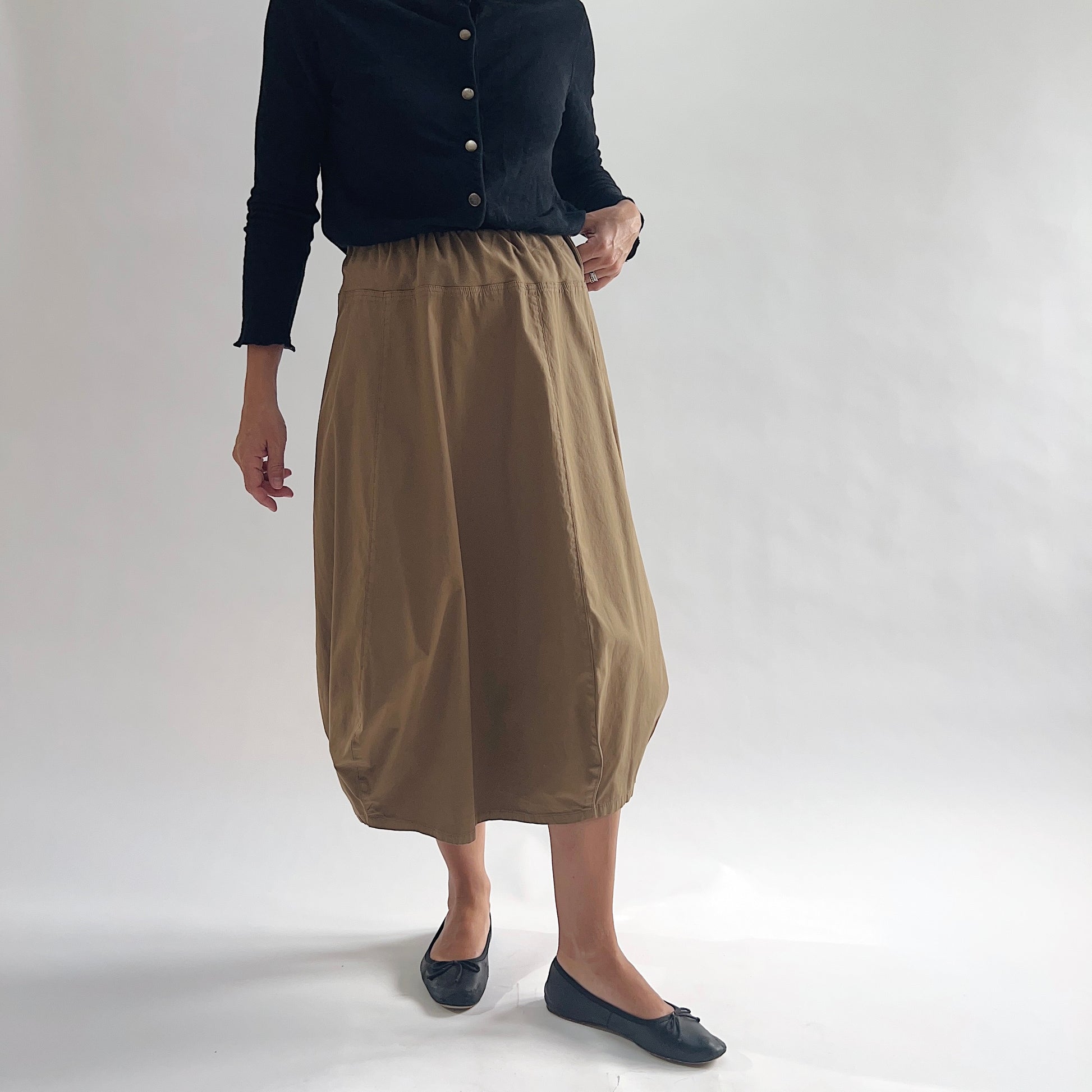Full front view of model wearing Eleven Stitch winter olive rib yoke skirt with arms at sides, showing elastic knit waistband, curved seam paneling, sculptural bubble hem, below-the-knee length, and warm khaki olive color, styled with black button-front cardigan and black ballet flats
