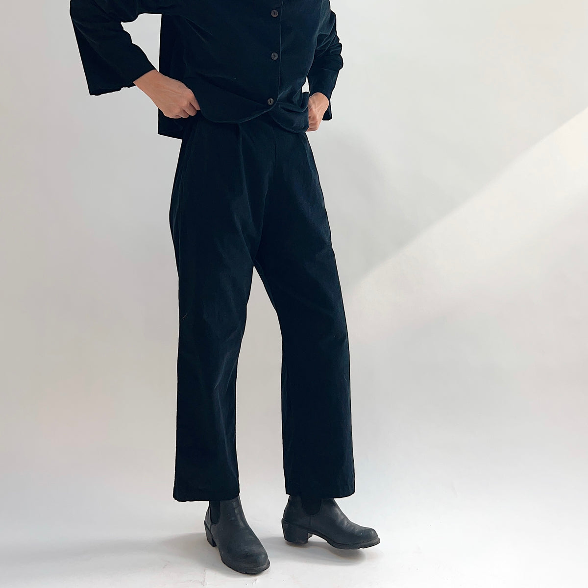 Model wearing the Bryn Walker Corduroy Pleated Pants in Black, cropped front view showing front pleats, flat waistband, straight leg silhouette, and black pinwheel corduroy, styled with matching Standing Collar Crop Jacket and black Chelsea boots
