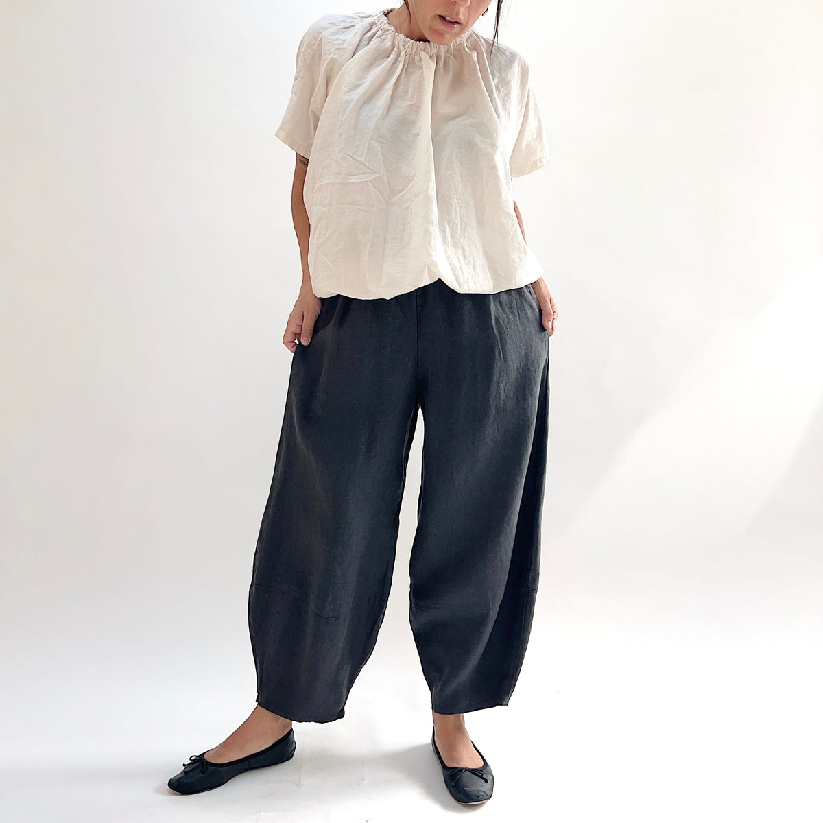 Model wearing the Bryn Walker Heavy Linen Oliver Pant in Moonlight, full front view highlighting barrel-leg cocoon silhouette, elastic waistband, and tapered ankle in deep moonlight charcoal heavy linen, styled with cream gathered-neck linen top and black ballet flats
