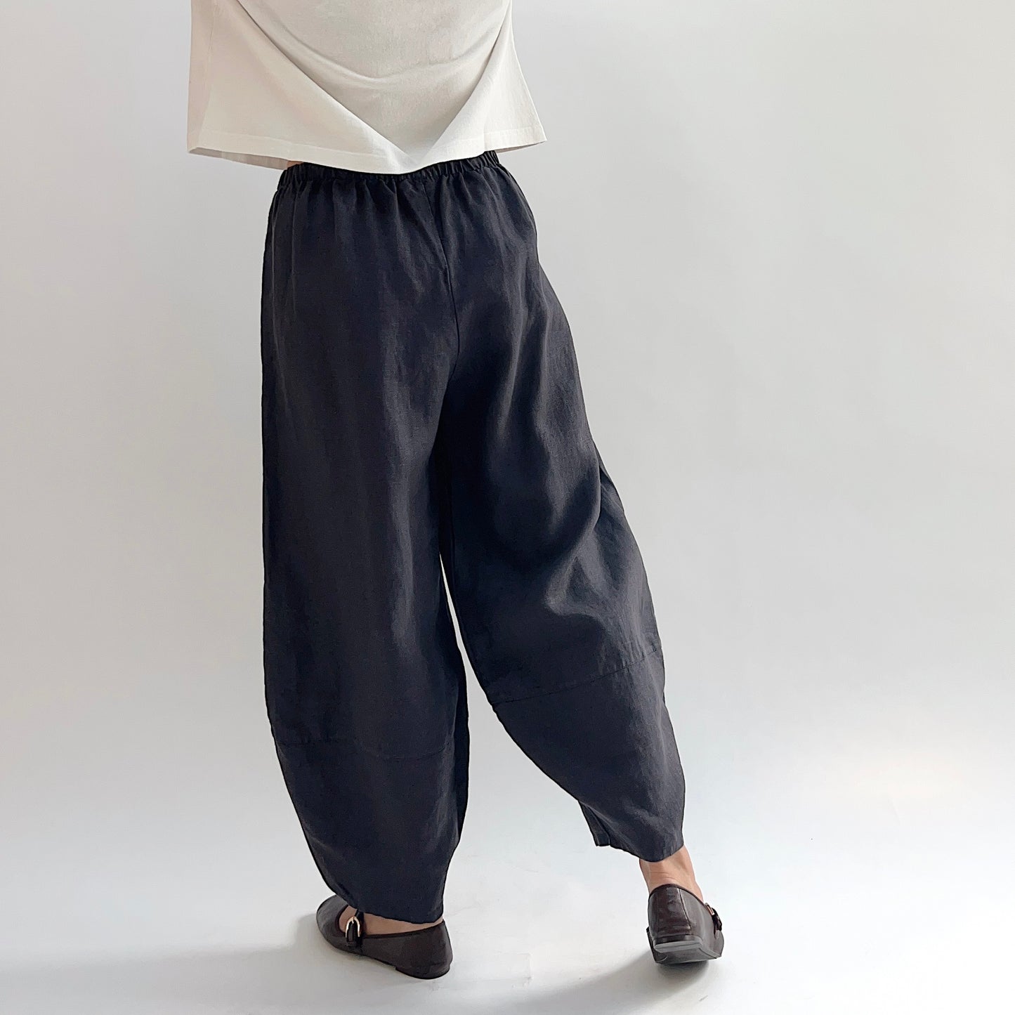 Back view of model wearing the Bryn Walker Heavy Linen Oliver Pant in Fig, showing elastic waistband, full barrel-leg drape, tapered ankle, and deep fig heavy linen texture, styled with cream top and brown Mary Jane flats
