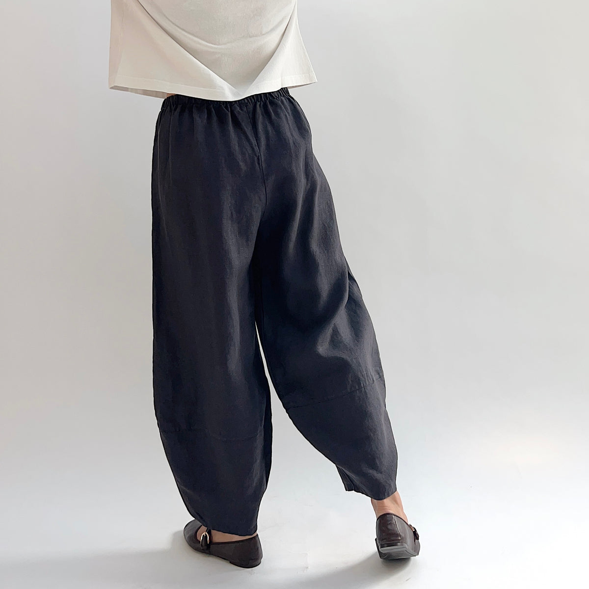 Back view of model wearing the Bryn Walker Heavy Linen Oliver Pant in Fig, showing elastic waistband, full barrel-leg drape, tapered ankle, and deep fig heavy linen texture, styled with cream top and brown Mary Jane flats
