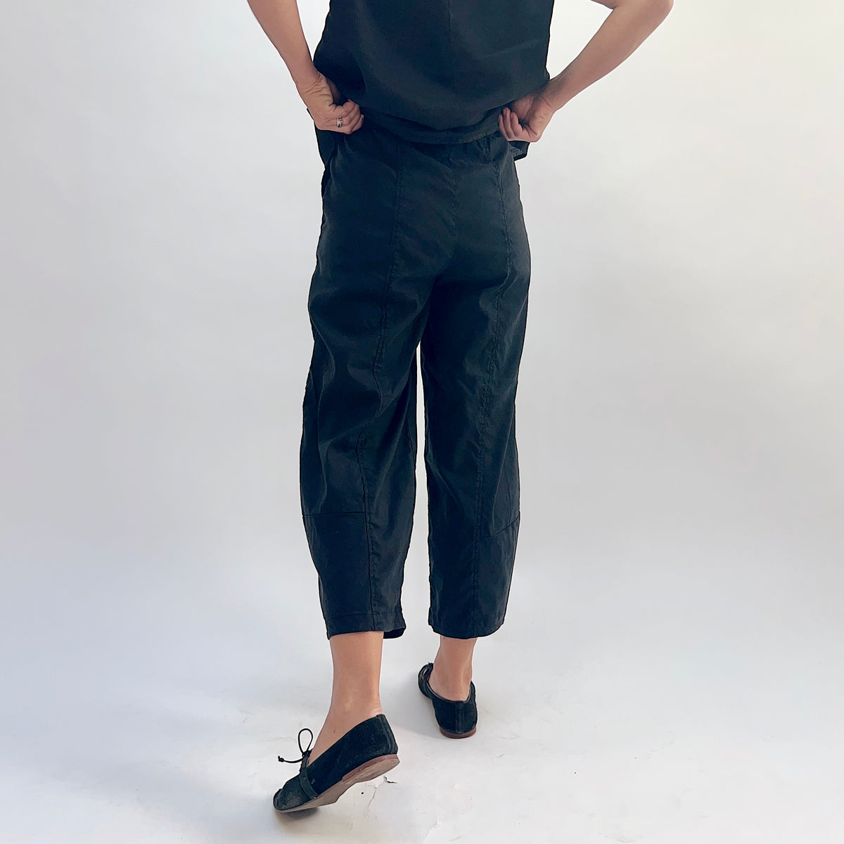Back view of black linen-tencel lantern pants showing curved seam detail down both legs, elastic waistband, and cropped ankle length, hands on hips, worn with a black linen top and black Mary Jane flats
