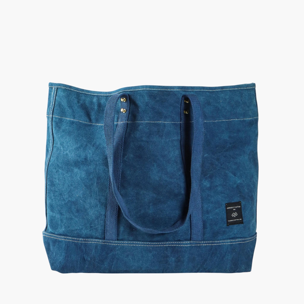 Indigo canvas tote with long blue webbing shoulder handles and a black Immodest Cotton label.

