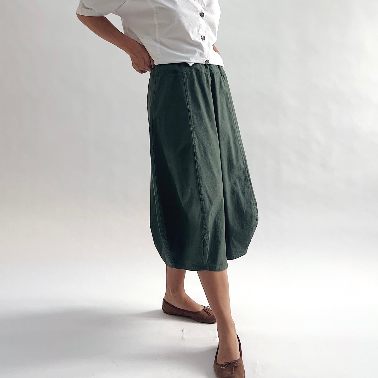 Three-quarter front view of model wearing Eleven Stitch konbu rib yoke skirt with hand on hip, showing elastic knit waistband, curved seam paneling, sculptural bubble hem, below-the-knee length, and deep forest green color, styled with white chalk camp shirt and brown ballet flats