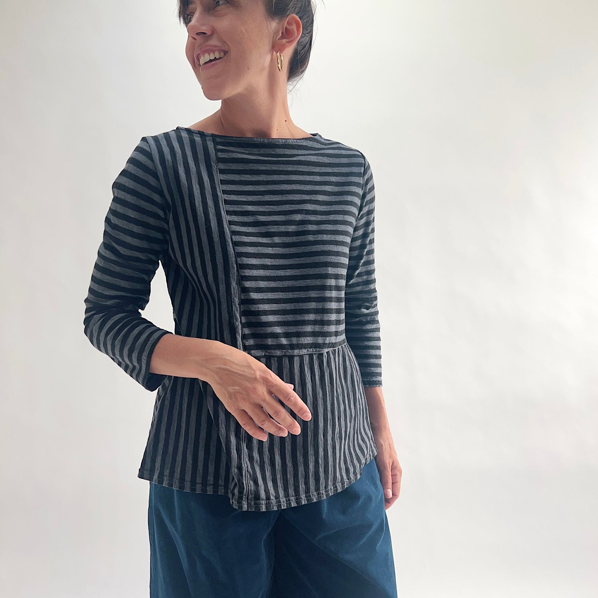 Three-quarter front view of smiling model wearing a charcoal and gray patchwork stripe A-line boatneck top, showing the mixed-direction stripe panel detail and gathered A-line hem, paired with dark blue wide-leg pants.
