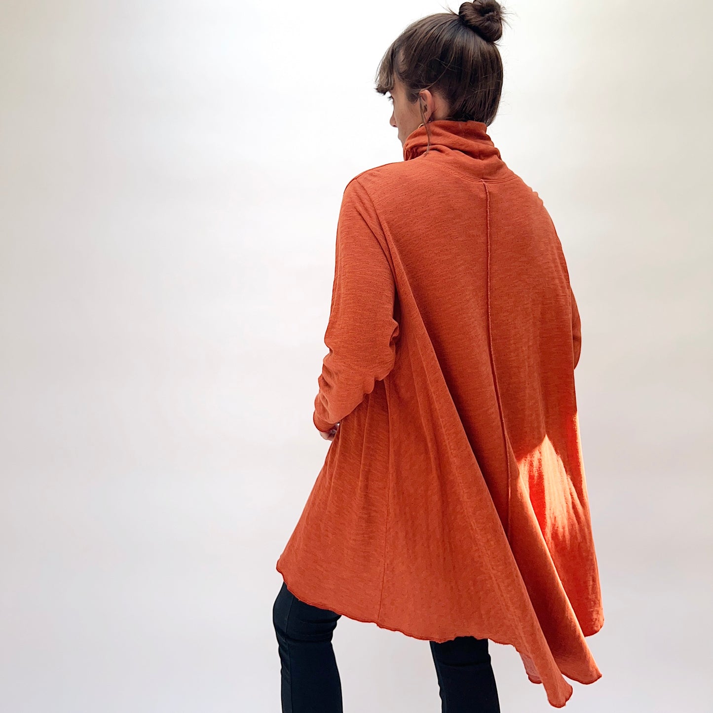 North Star Base Double Cotton Hi Low Turtleneck Top in Raw Sienna, back view on model, showing center back seam, wide A-line silhouette, turtleneck collar, long sleeves, and flowing hi-low raw hem, paired with dark jeans
