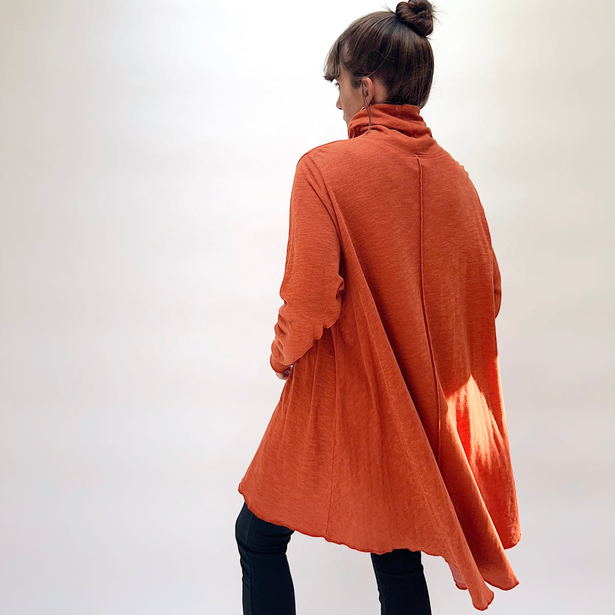 North Star Base Double Cotton Hi Low Turtleneck Top in Raw Sienna, back view on model, showing center back seam, wide A-line silhouette, turtleneck collar, long sleeves, and flowing hi-low raw hem, paired with dark jeans
