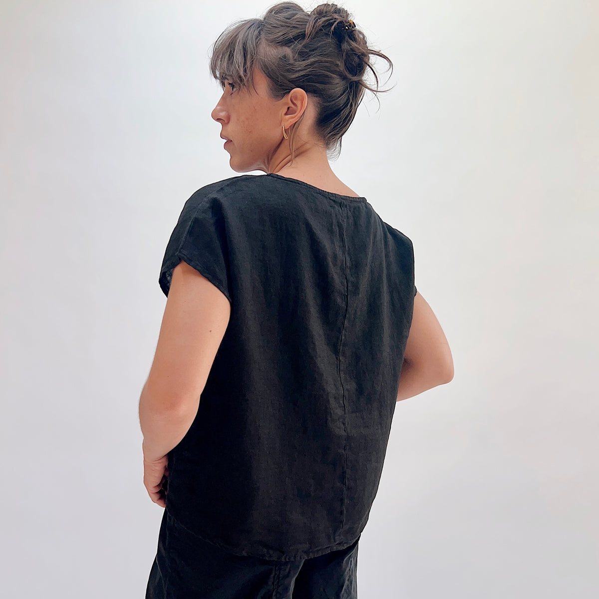 Back view of black linen high-low tee showing center back seam, boatneck, and cap sleeves, hair up with gold hoop earrings, styled with black linen lantern pants
