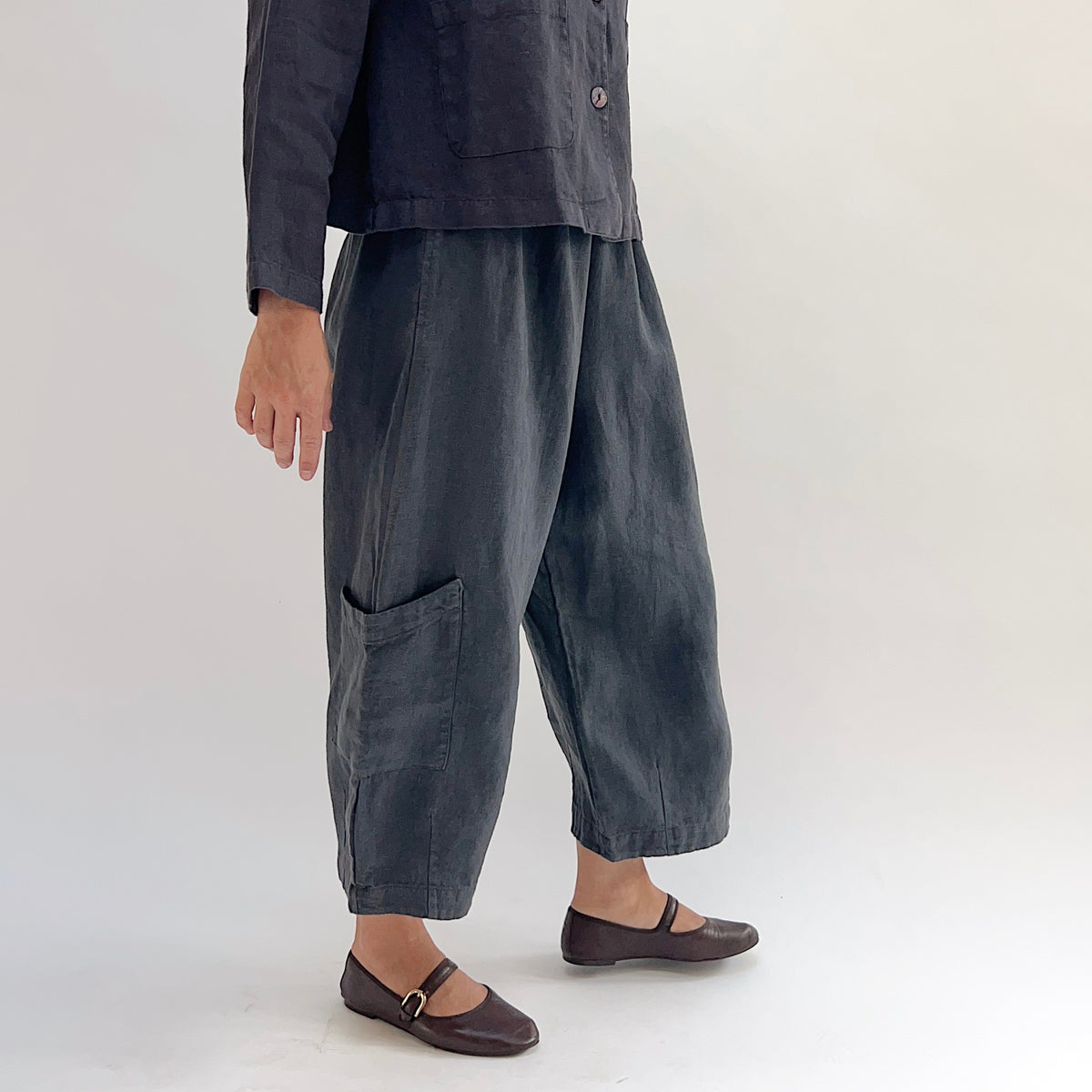 Model wearing the Bryn Walker Heavy Linen Casbah Pant in Moonlight, side view showing elastic waistband, hip pocket, barrel-tapered leg, large lower-leg patch pocket, cropped ankle length, and deep moonlight heavyweight linen, styled with matching linen jacket and brown Mary Jane flats
