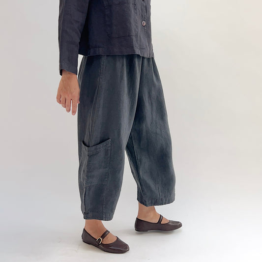 Model wearing the Bryn Walker Heavy Linen Casbah Pant in Moonlight, side view showing elastic waistband, hip pocket, barrel-tapered leg, large lower-leg patch pocket, cropped ankle length, and deep moonlight heavyweight linen, styled with matching linen jacket and brown Mary Jane flats
