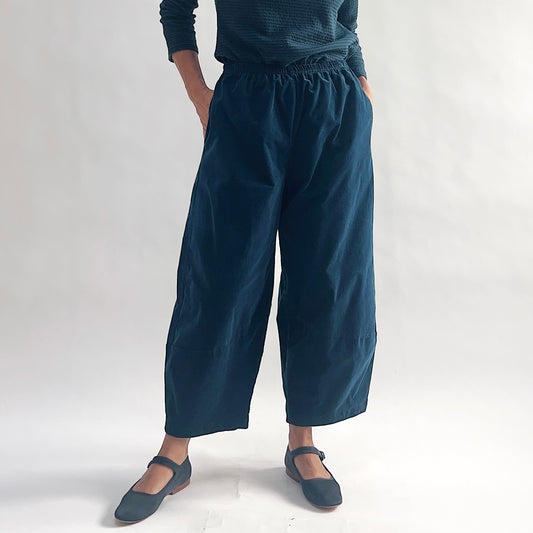 Front view of model wearing deep teal mini cord lantern pants with hands in side pockets, showing the gathered elastic waist, wide rounded silhouette, and tapered ankle, styled with a matching teal textured long-sleeve top and teal mary jane flats against a white background.
