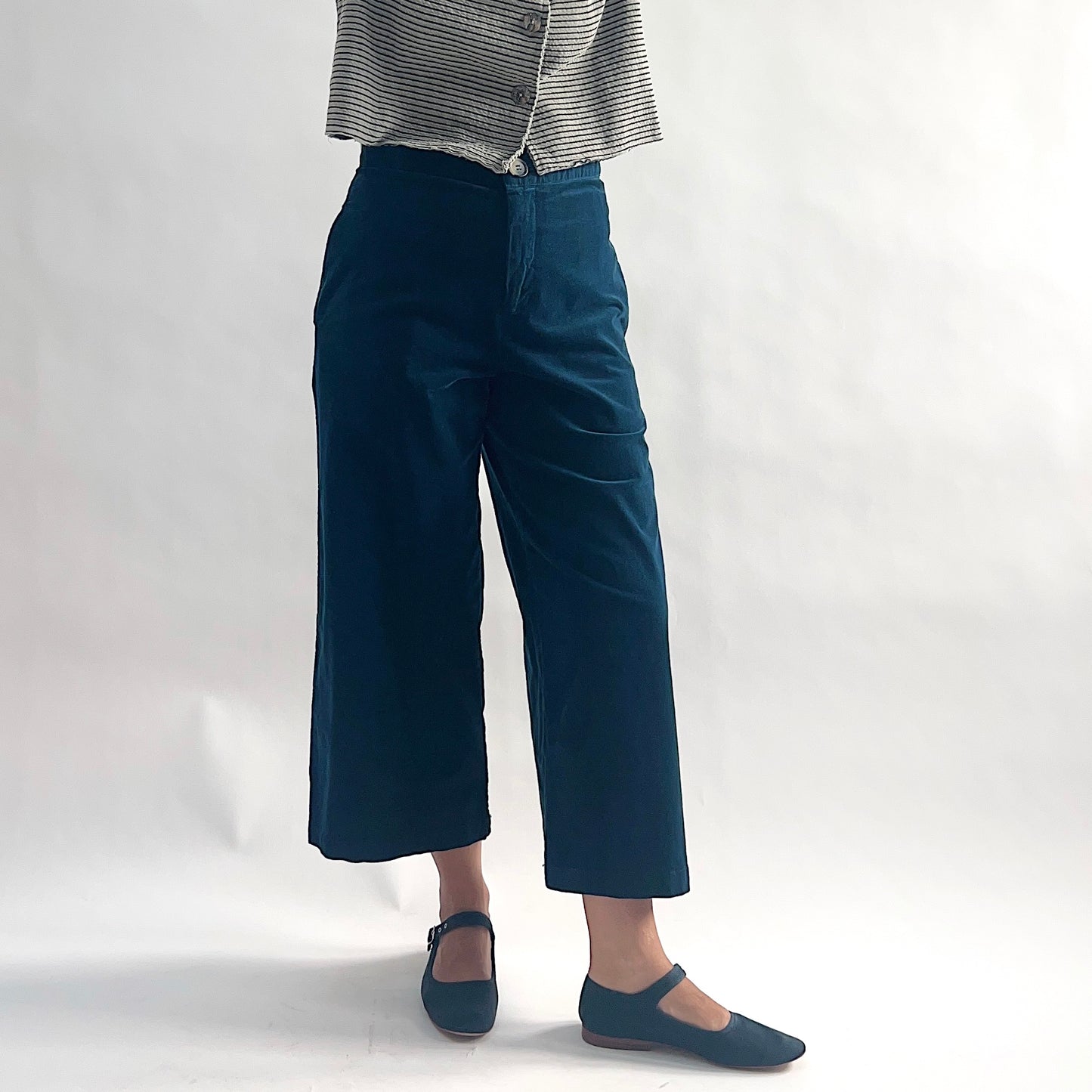 Three-quarter side view of model wearing deep teal mini cord straight crop trousers, showing the flat front, side seam pocket, wide straight leg, and cropped hem, styled with the matching white and black stripe asymmetrical cardigan and teal mary jane flats against a white background.
