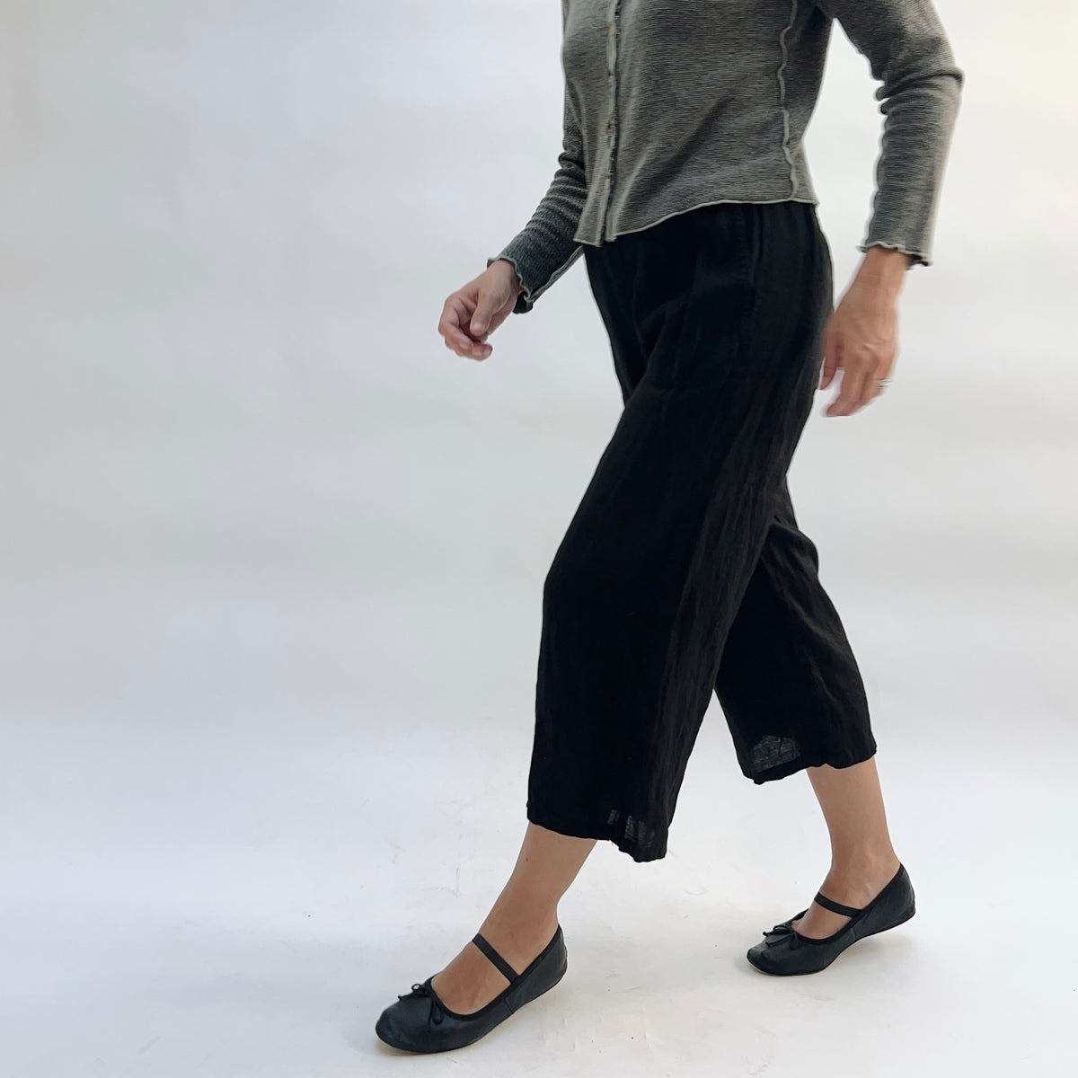 Cut Loose Hanky Linen Easy Crop Flood Pant in Black, black linen wide-leg crop pant walking forward, styled with a grey button-front cardigan and black Mary Jane flats — angled side view showing silhouette and hem vent in motion
