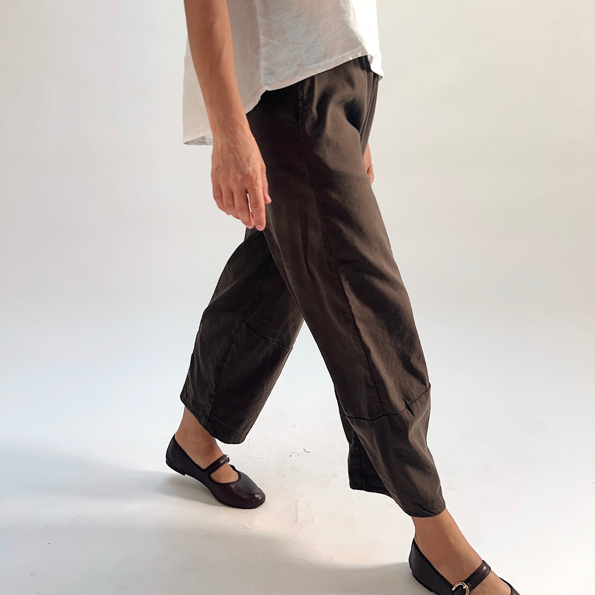 Walking side-angle view of walnut linen-tencel lantern pants showing wide barrel-leg silhouette in motion, curved ankle hem seam, styled with a white linen top and dark Mary Jane flats
