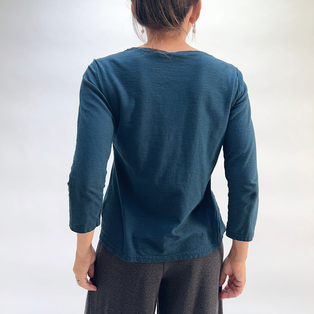 Model wearing the Cut Loose 3/4 Sleeve Boatneck Top in Etan, back view, showing clean back seaming, side vents, and relaxed fit of deep teal cotton-linen boatneck top with 3/4 sleeves, styled with black textured pants
