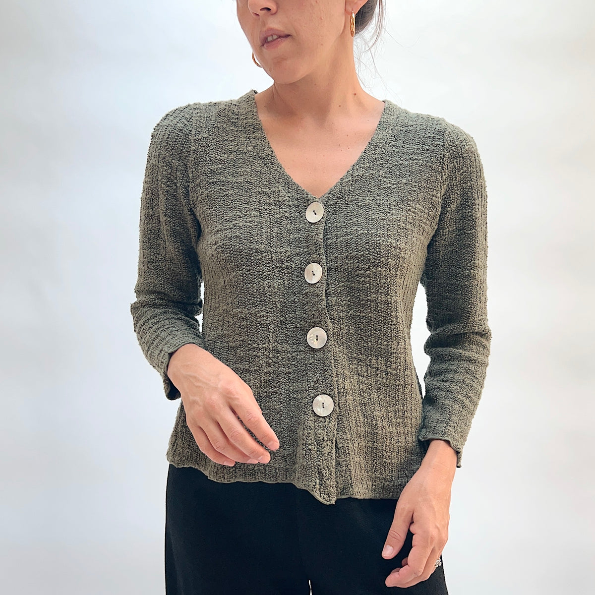 Model wearing the Cut Loose Puff Sleeve Cardigan in Kelp, full-length front view, showing V-neckline, four large natural shell buttons, slightly fitted silhouette, and scalloped lettuce hem of olive green marled knit cardigan, styled with black pants
