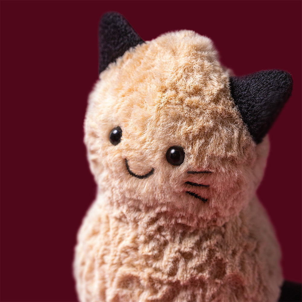 Close-up of the Jellycat Amuseables Peanut Cat face against a deep red background, showing its golden fluffy texture, black button eyes, embroidered smile, and dark ears