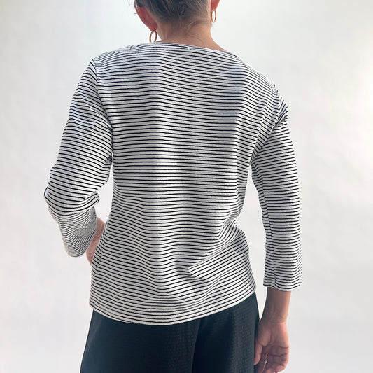 Back view of model wearing a white and black horizontal stripe boatneck tee with 3/4 sleeves, hand on hip, showing the wide boatneck, curved hem, and crinkle-texture fabric detail, styled with black wide-leg pants.
