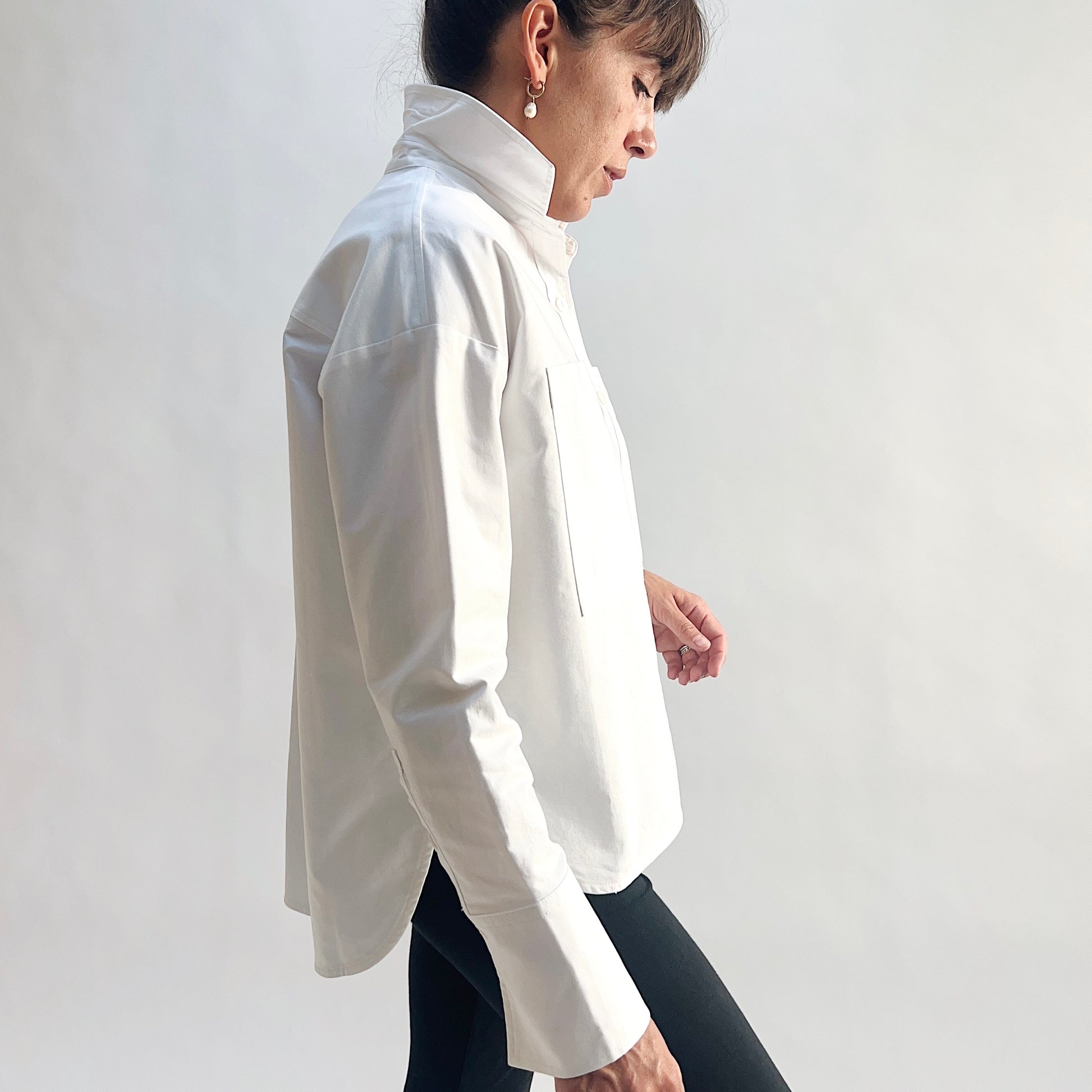 Side profile view of model walking in a white cotton oxford boyfriend shirt, showing the relaxed oversized fit, longer curved shirttail, side hem slit, and wide barrel cuff, styled with black slim pants against a white background.
