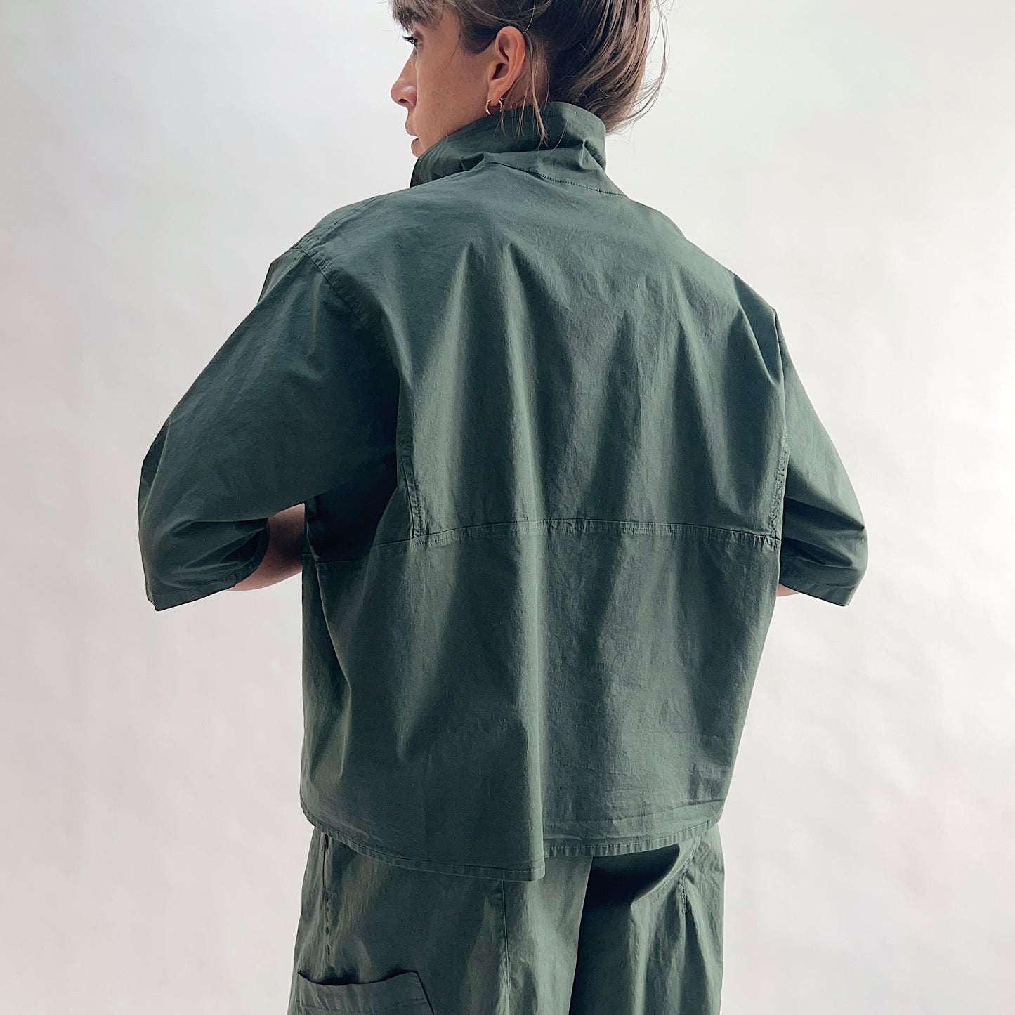 Back view of model wearing Eleven Stitch konbu camp shirt with hands on hips, showing rounded collar, back yoke seam, short sleeves, slightly cropped boxy silhouette, and deep forest green color, styled with matching konbu snap front pant