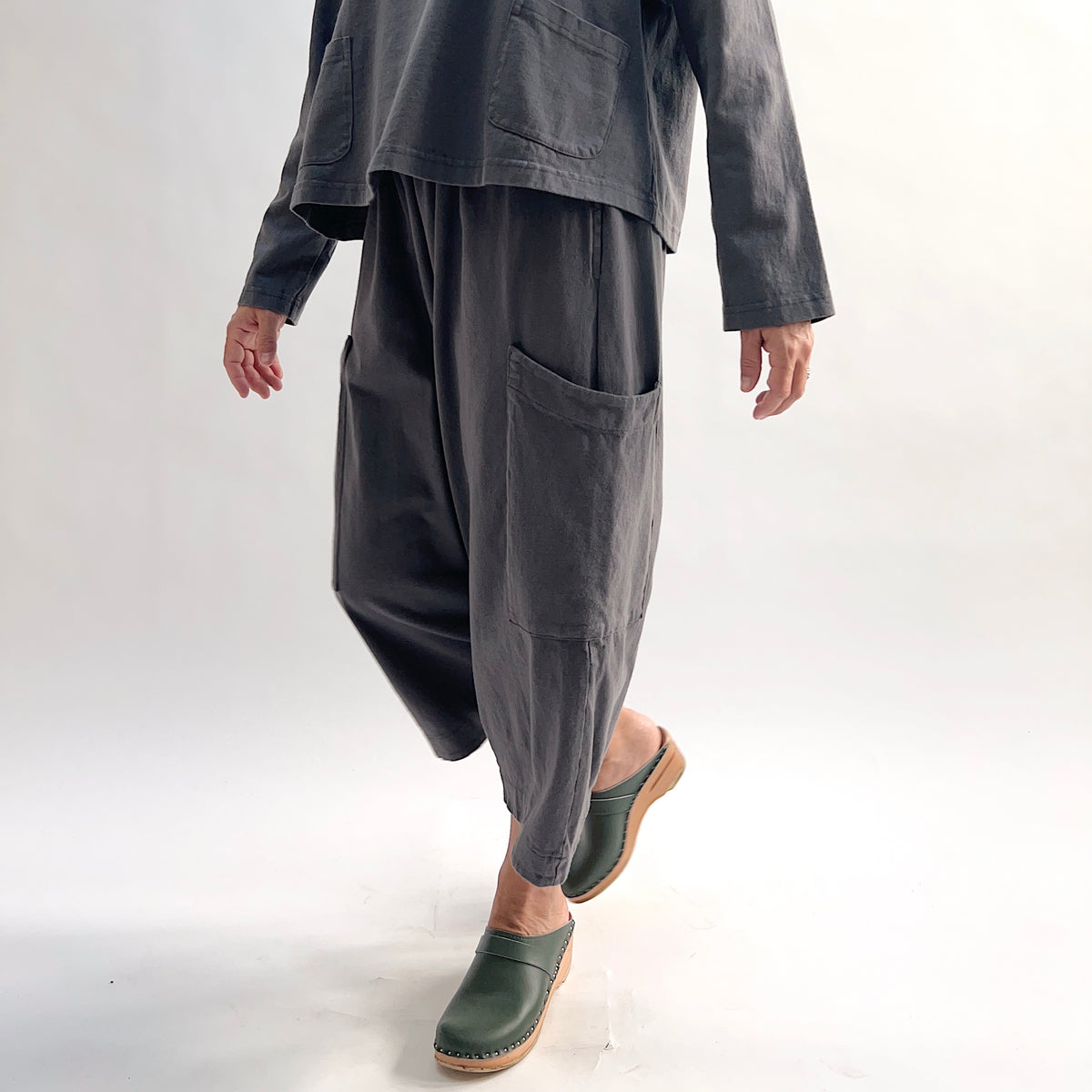 Model walking in the Bryn Walker Cotton Pasha Pant in Moonlight, side view showing movement in wide-leg silhouette, thigh patch pockets, elastic gathered waistband, and soft blue-grey cotton drape, styled with matching top and green wedge clogs
