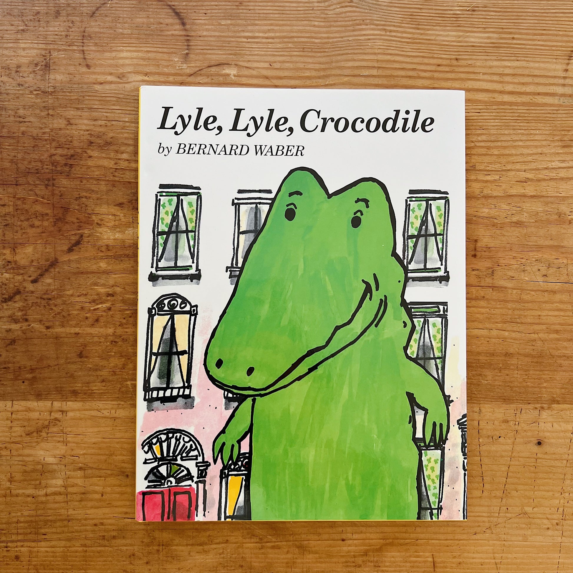 Front cover of Lyle, Lyle, Crocodile hardcover by Bernard Waber, displayed flat on a warm wood surface. White background with the title in large bold black serif lettering at the top and "by BERNARD WABER" in smaller caps below it. The central illustration shows Lyle — a large, cheerful bright green crocodile — standing close-up in front of a pink New York City brownstone façade, with arched windows, iron railings, and a red front door with a yellow lamppost visible behind him. Lyle's expression is gentle a