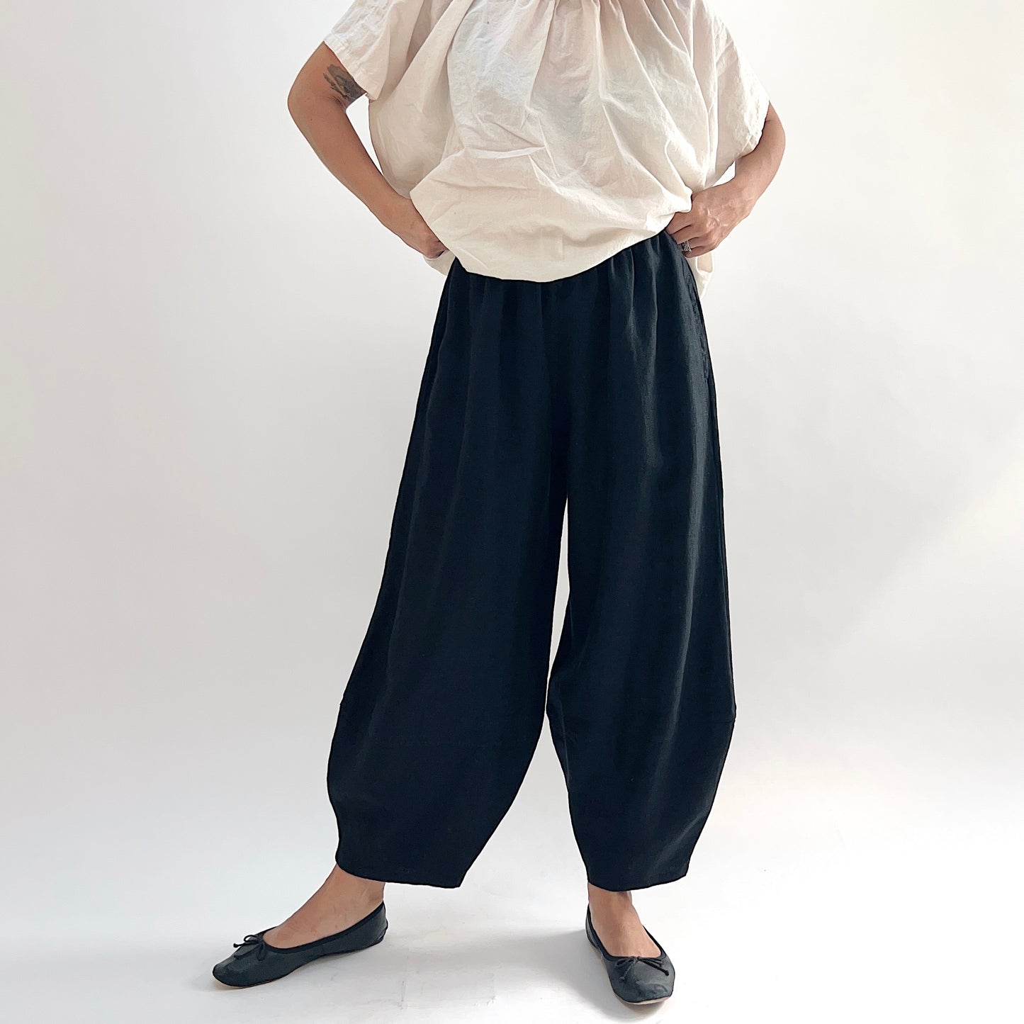 Model with hands on hips wearing the Bryn Walker Heavy Linen Oliver Pant in Black, full front view showing barrel-leg silhouette, elastic waistband, and tapered ankle in black heavy linen, styled with cream gathered linen top and black ballet flats
