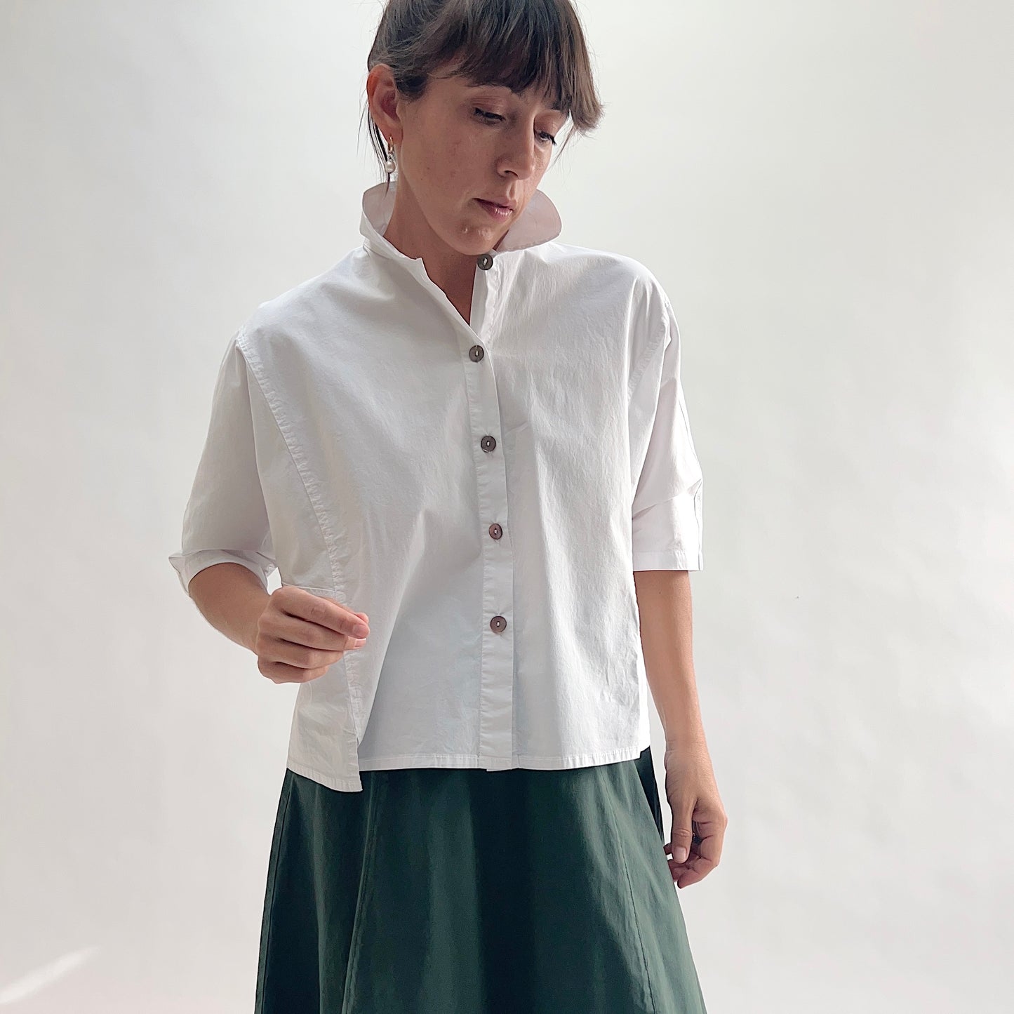 Three-quarter front view of model wearing Eleven Stitch chalk camp shirt looking down, showing stand collar, dark shell buttons, short sleeves, slightly cropped boxy fit, side slits, and white color, styled with forest green wide-leg skirt

