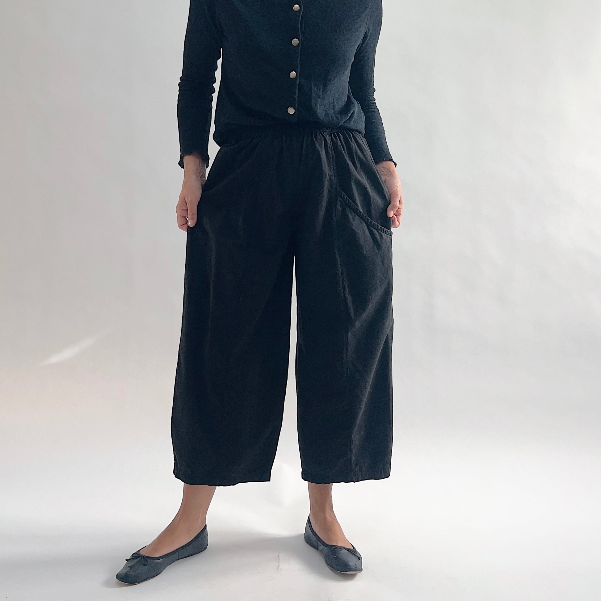 Close front view of model wearing Eleven Stitch black one pocket pant, showing elastic waistband, wide cropped leg, large curved side pocket, gathered elastic hem, and black color, styled with black long sleeve button front top and black ballet flats