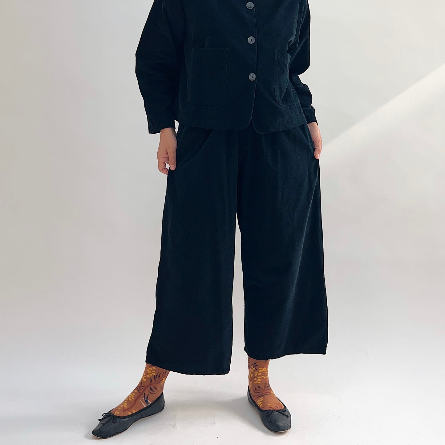 Model wearing the Bryn Walker Corduroy Flood Pants in Black, full front view showing elastic waistband, relaxed wide-leg flood silhouette, and black pinwheel corduroy, paired with matching Standing Collar Crop Jacket, printed socks, and black ballet flats

