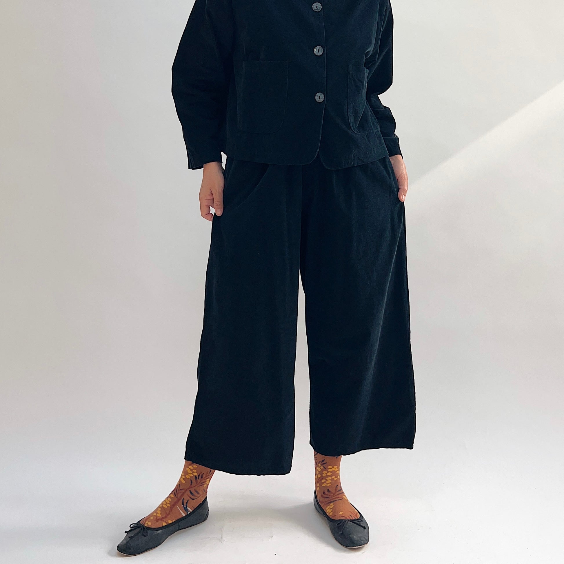 Model wearing the Bryn Walker Corduroy Flood Pants in Black, full front view showing elastic waistband, relaxed wide-leg flood silhouette, and black pinwheel corduroy, paired with matching Standing Collar Crop Jacket, printed socks, and black ballet flats
