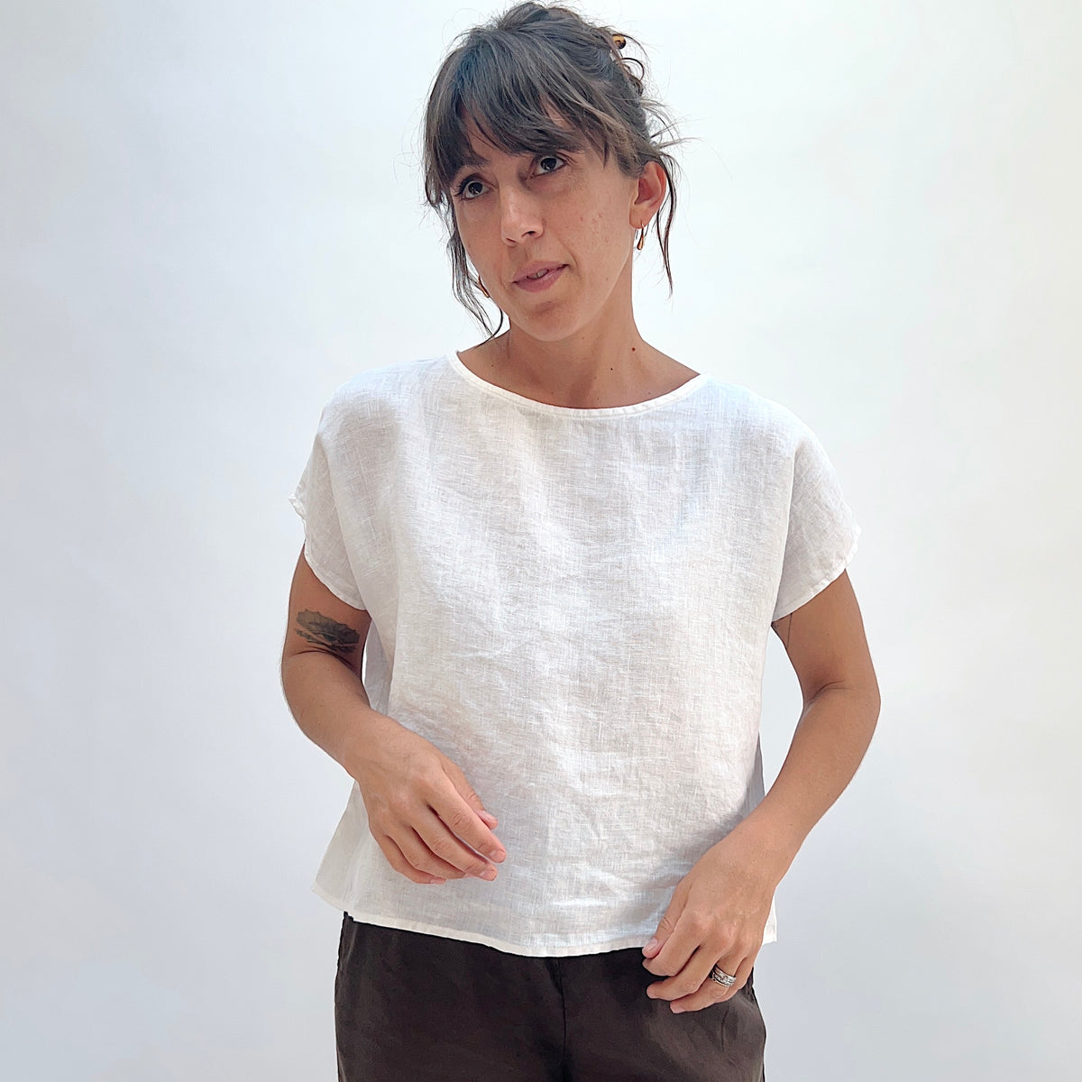 Front view of white linen high-low tee with boatneck and cap sleeves, showing relaxed boxy fit, paired with dark trousers
