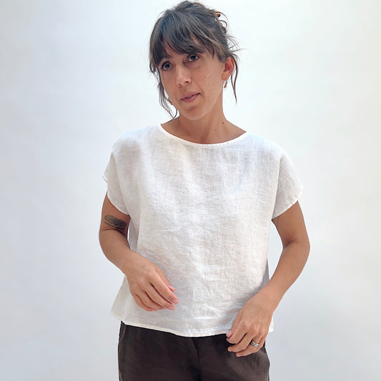 Front view of white linen high-low tee with boatneck and cap sleeves, showing relaxed boxy fit, paired with dark trousers
