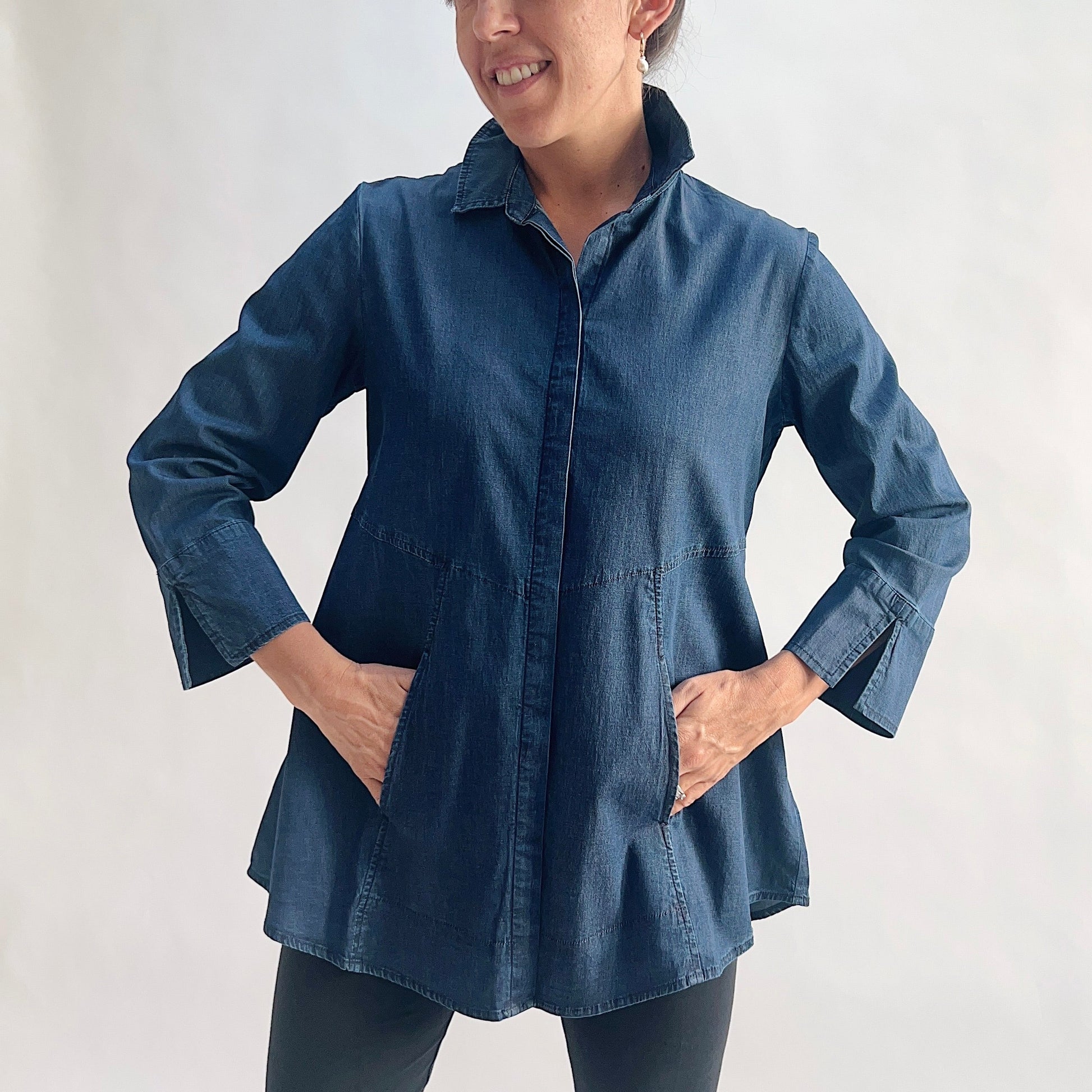 Full front view of model wearing a lightweight dark indigo denim tunic shirt with hands in built-in side pockets, showing the hidden front placket, empire seam, wide flared A-line hem, and slit cuffs, styled with black slim pants against a white background.
