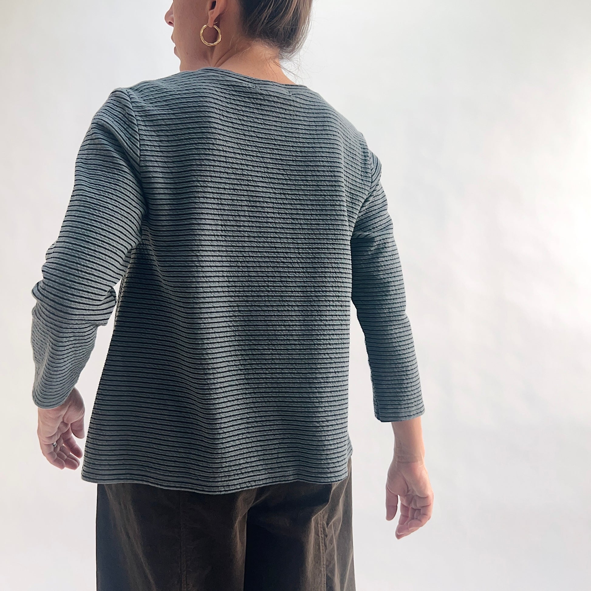 Back view of model wearing an iron charcoal and black tonal textured stripe crewneck top with 3/4 sleeves, showing the curved hem, side vents, and crinkle-texture fabric, styled with dark olive wide-leg pants against a white background.
