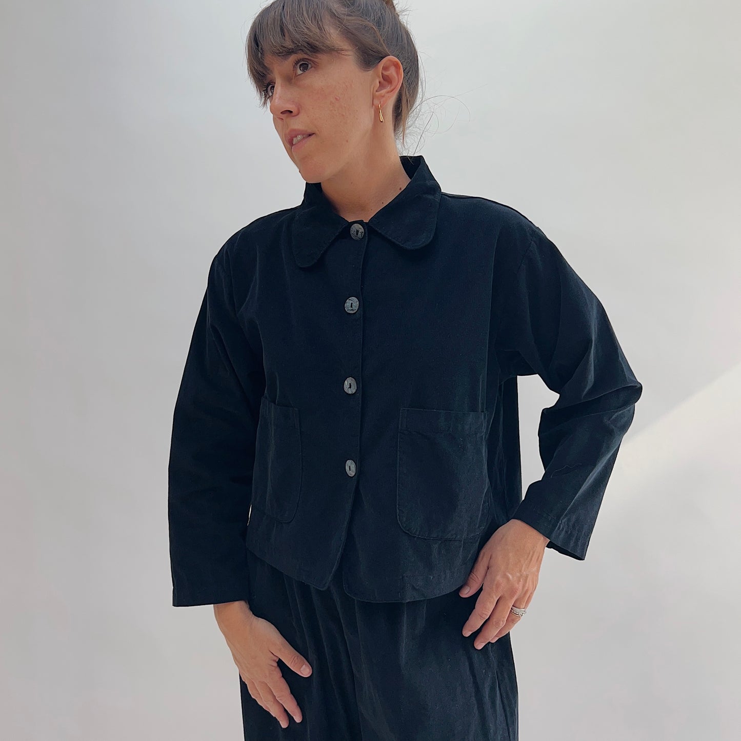 Model with hands on hips wearing the Bryn Walker Corduroy Audrey Jacket in Black, front view showing rounded peter-pan collar, shell buttons, and patch pockets, paired with black trousers
