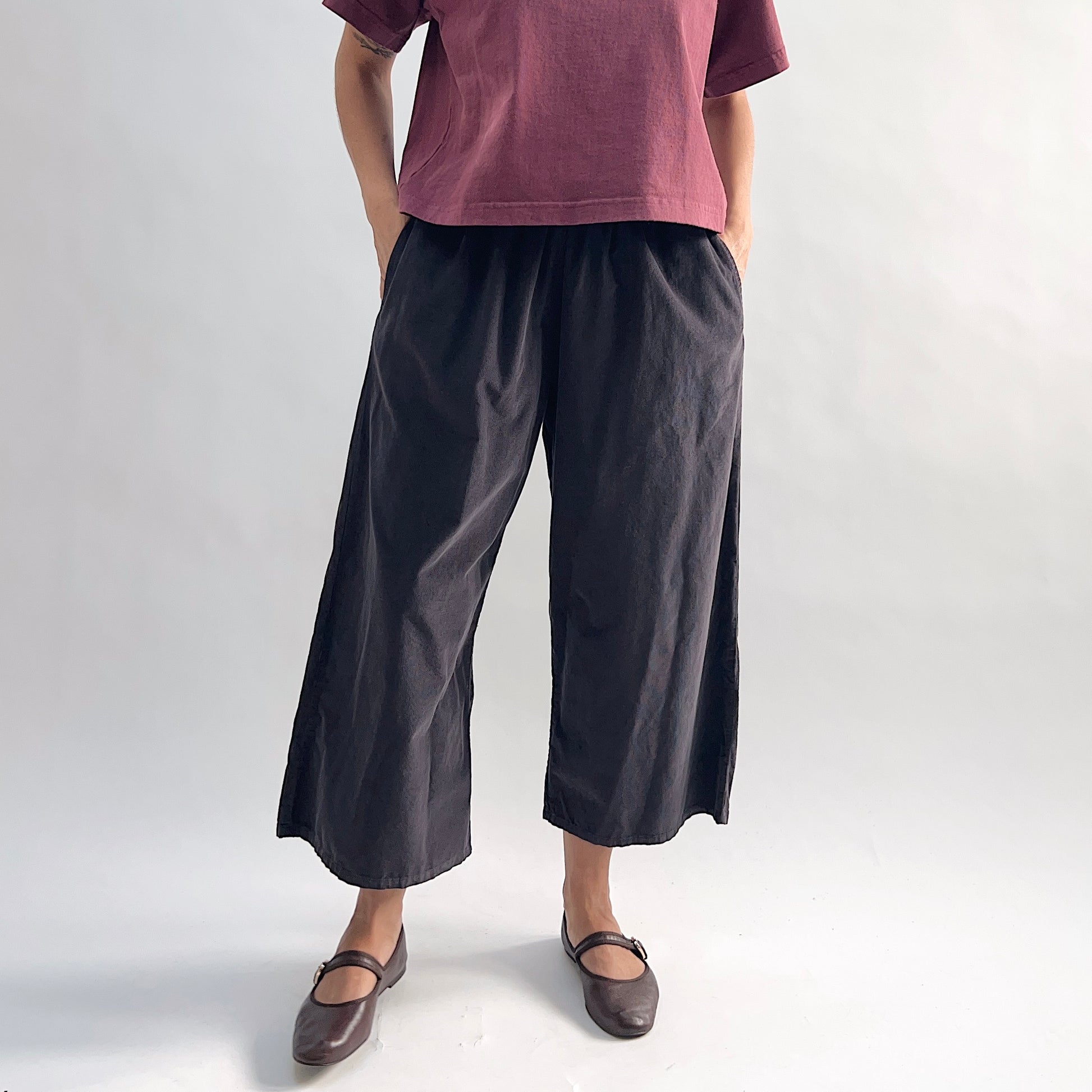 Model with hands in pockets wearing the Bryn Walker Corduroy Flood Pants in Fig, full front view showing elastic gathered waistband, wide culotte-style leg, mid-calf flood length, and deep fig pinwheel corduroy, styled with burgundy Double Cotton Tee and brown Mary Jane flats
