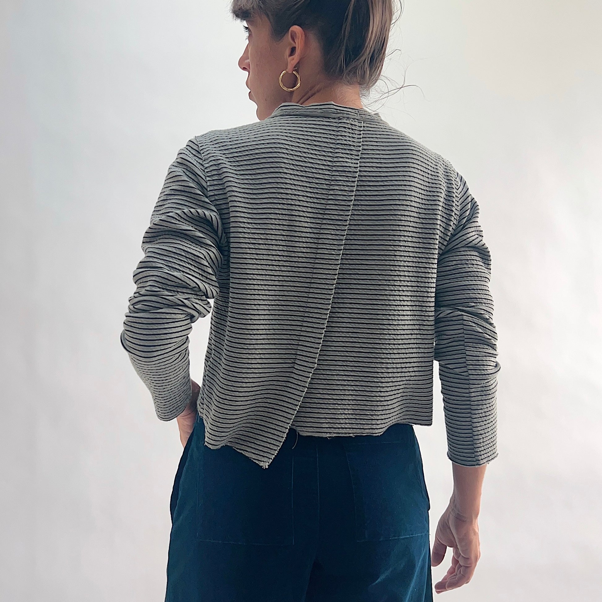 Back view of model wearing a squirrel and black fine horizontal stripe cropped cardigan with long sleeves and asymmetrical hem, hand in pocket, showing the crinkle-texture fabric and center back seam, styled with dark teal wide-leg pants against a white background.

