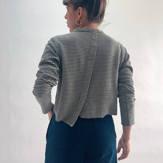Back view of model wearing a squirrel and black fine horizontal stripe cropped cardigan with long sleeves and asymmetrical hem, hand in pocket, showing the crinkle-texture fabric and center back seam, styled with dark teal wide-leg pants against a white background.
