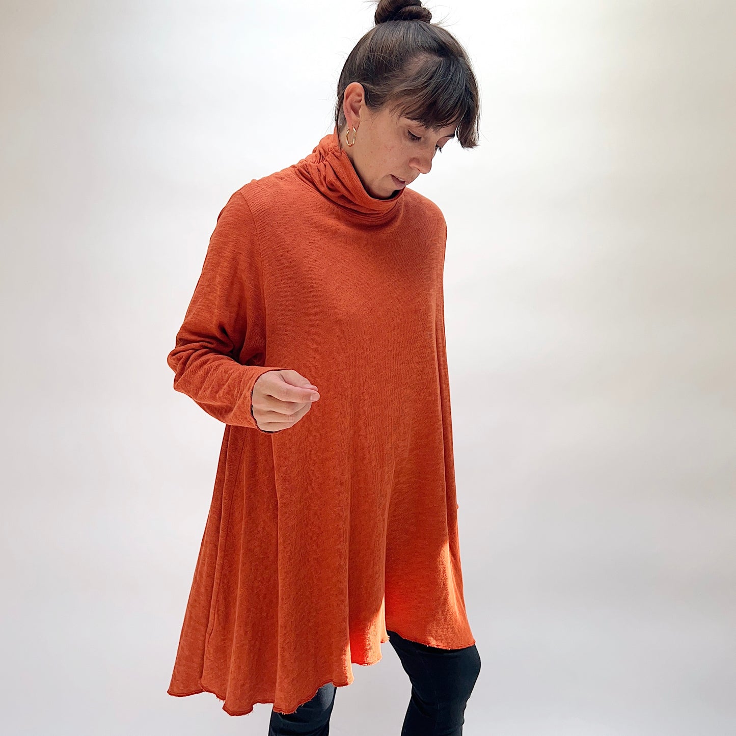 North Star Base Double Cotton Hi Low Turtleneck Top in Raw Sienna, full front view on model looking down, showing draped turtleneck collar, relaxed wide body, double-layered raw sienna fabric, and sweeping hi-low hem with raw edge, paired with dark jeans
