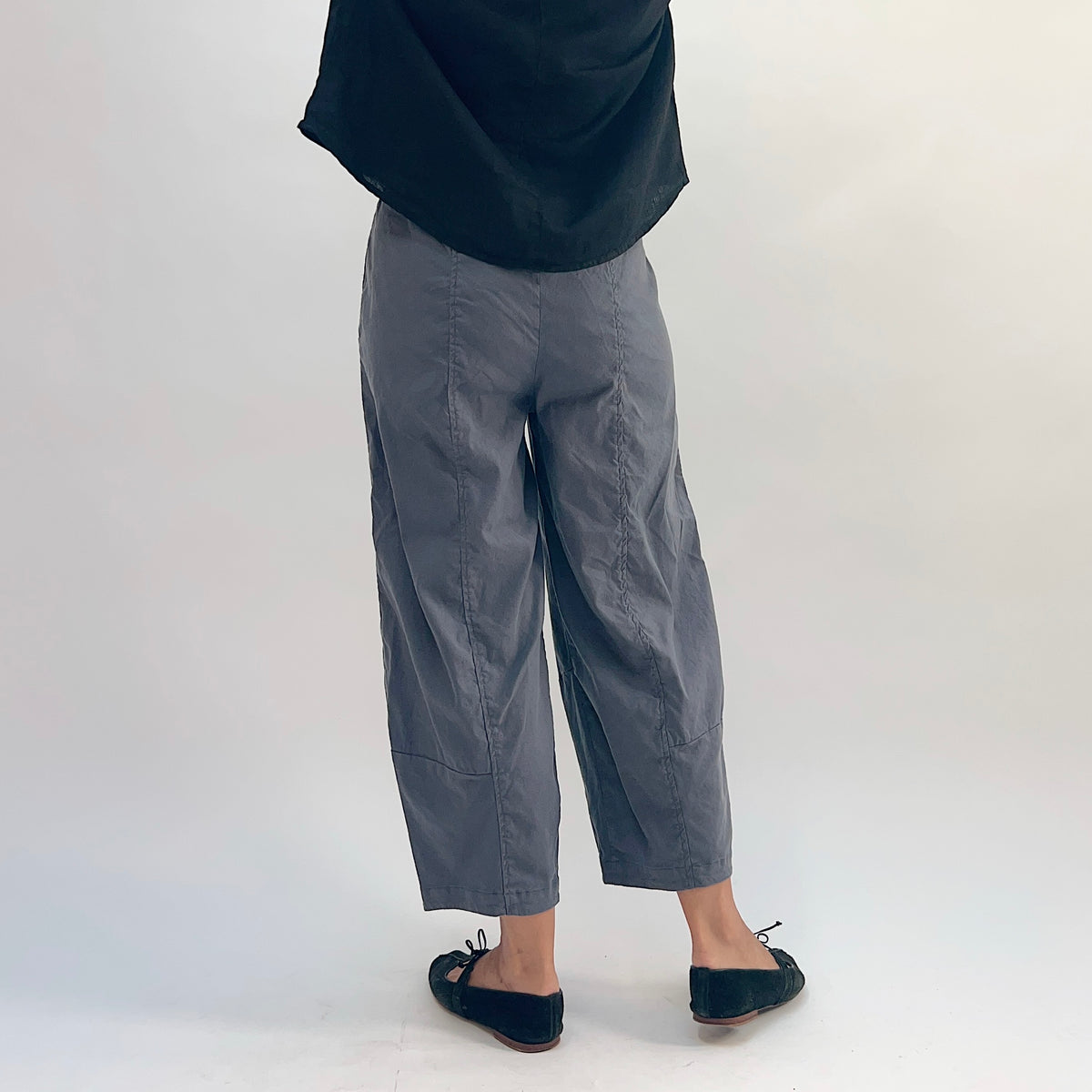 Back view of pewter linen-tencel lantern pants showing curved seam detail down both legs, wide barrel-leg silhouette, elastic waistband, and ankle-length hem, worn with a black linen top and black bow-tie ballet flats
