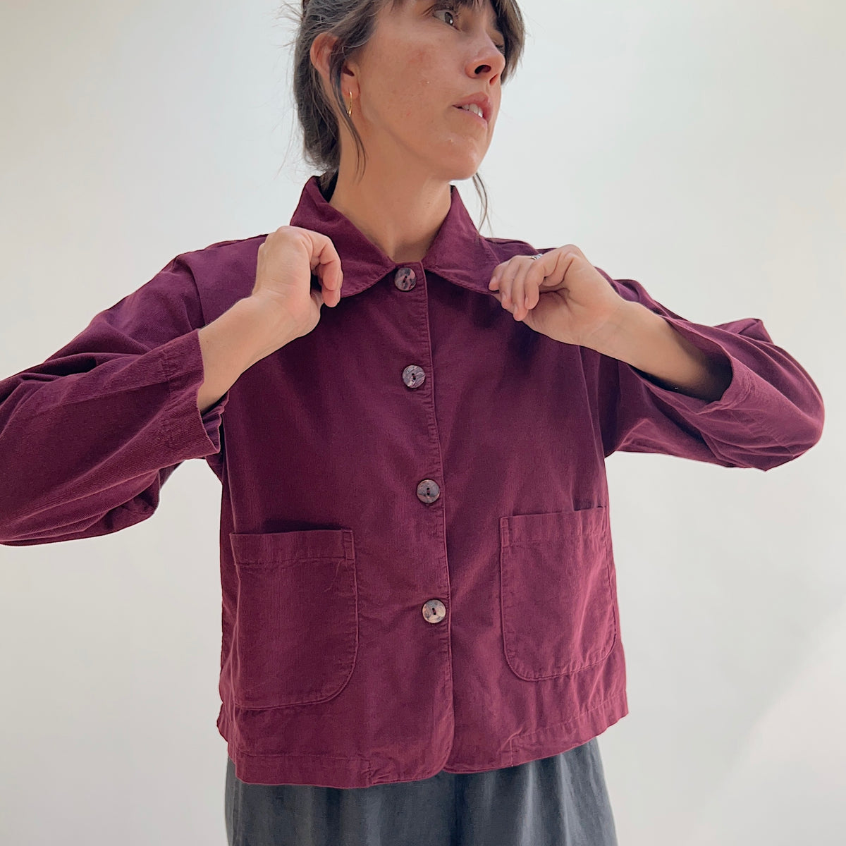 Model adjusting the collar of the Bryn Walker Corduroy Audrey Jacket in Rhone, close-up front view highlighting rounded collar, shell buttons, patch pockets, and burgundy pinwheel corduroy texture

