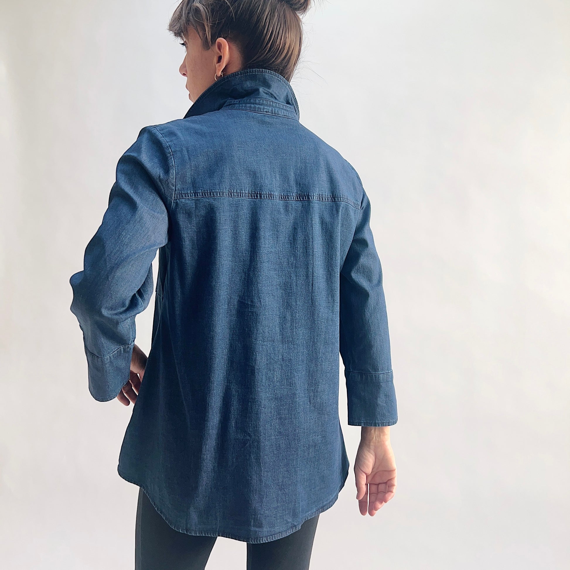 Back view of model wearing a lightweight dark indigo denim tunic shirt, showing the point collar, back yoke seam, 3/4 sleeves with slit cuffs, and curved shirttail hem, styled with black slim pants against a white background.
