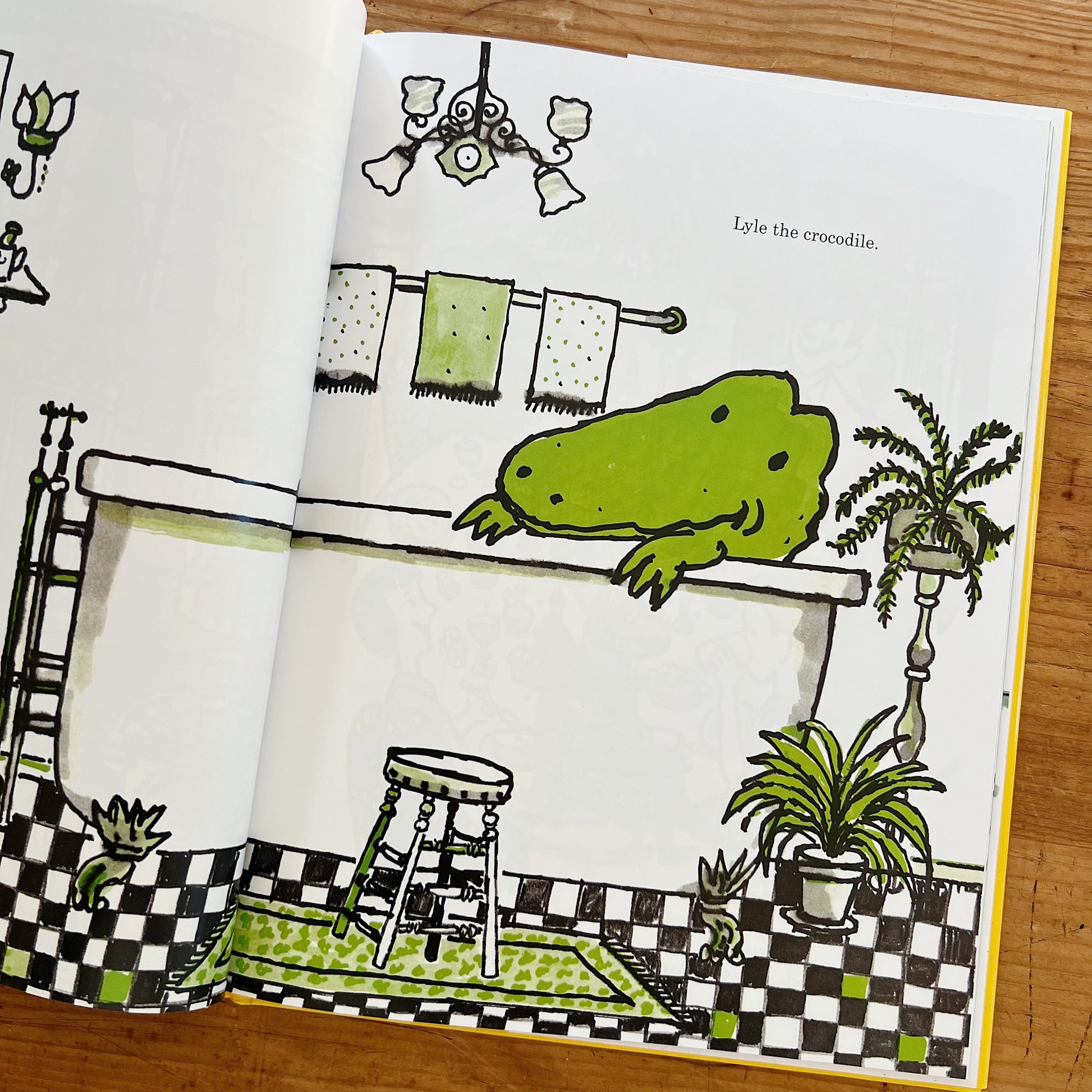 Open interior spread of Lyle, Lyle, Crocodile displayed on a warm wood surface, showing the introductory bathroom illustration across both pages. Lyle — a large bright green crocodile — is draped contentedly over the edge of a white claw-foot bathtub, his front claws hanging over the rim. The bathroom has black-and-white checkerboard tile floors, a patterned green bath mat, a wooden step stool, hanging towels on a wall rack, potted ferns on a pedestal, and an ornate black chandelier above. A small cat and c