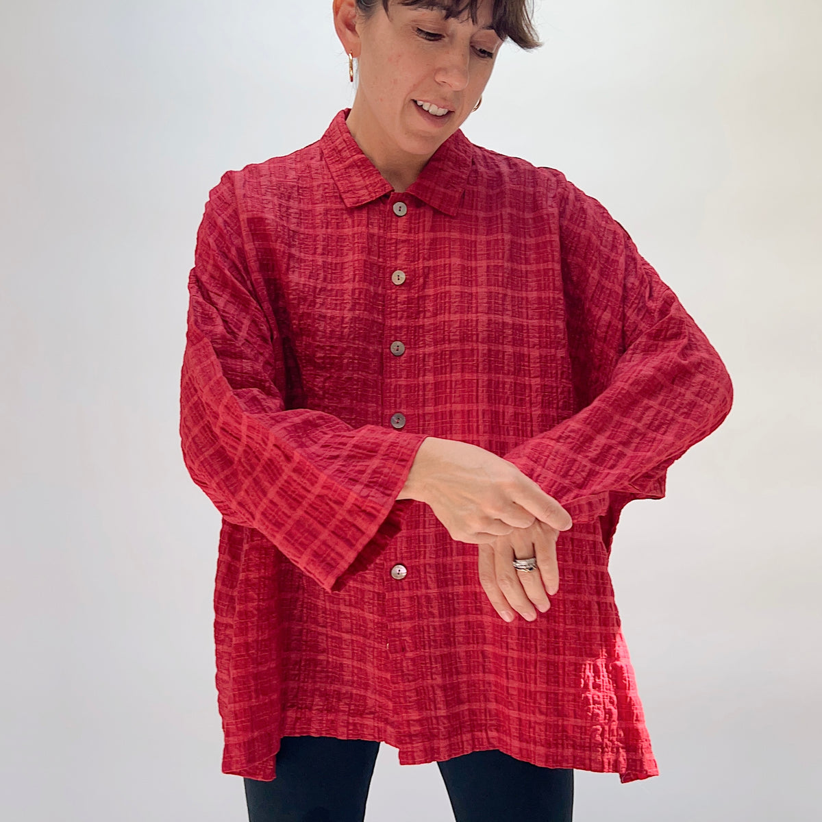 Eleven Stitch Big Shirt in Stripe Crinkle Ruby, front view on smiling model adjusting cuffs, showing point collar, five wood button closure, crinkle stripe texture in ruby, and relaxed oversized fit, paired with dark slim trousers
