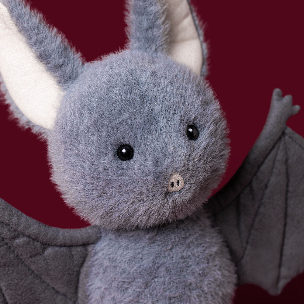 Close-up of the Jellycat Broox Bat face against a deep red background, showing soft blue-grey fur, large cream-lined ears, shiny black hard eyes, and a small pink nose
